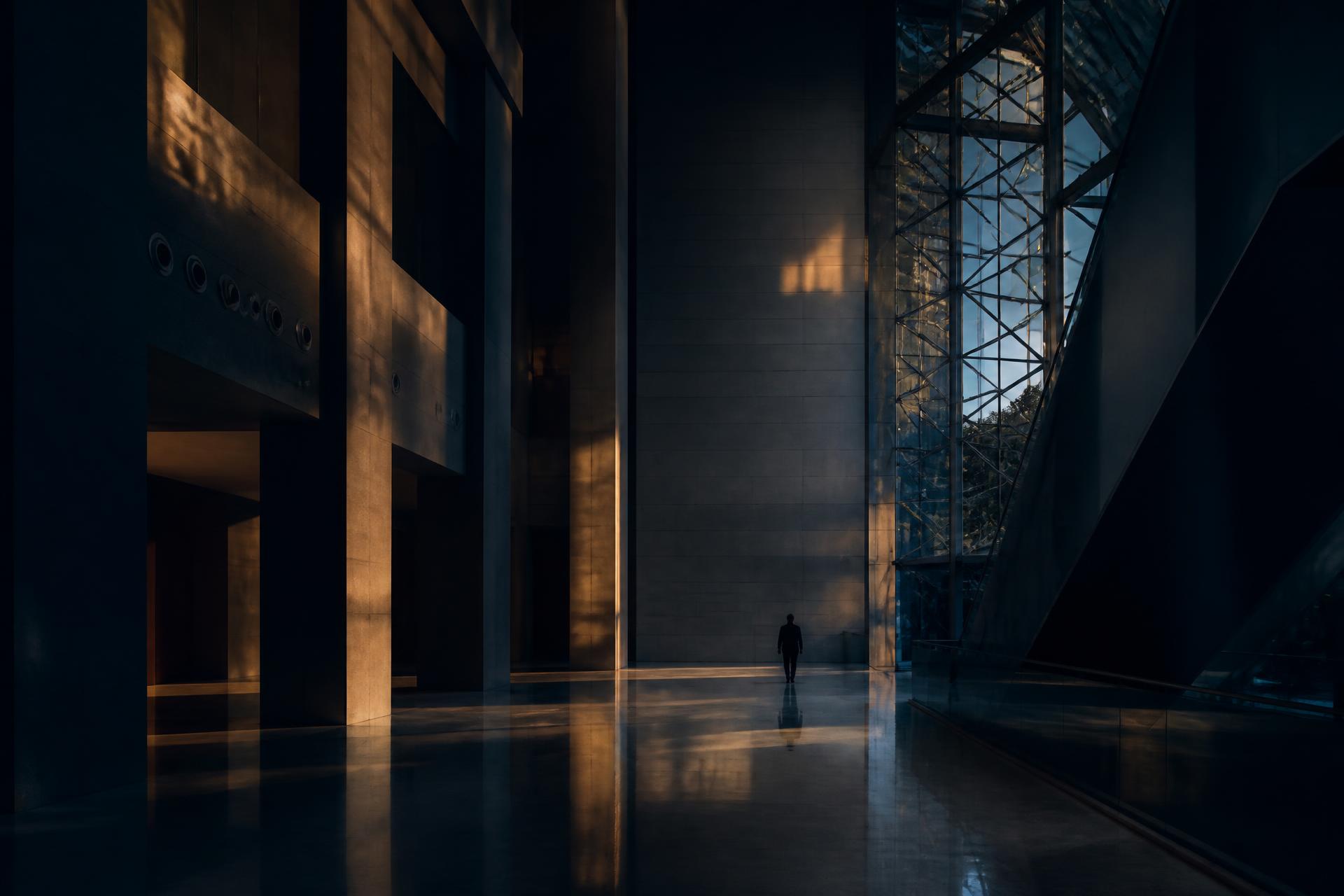 The quiet atrium of an institutional building at dusk, with a single figure crossing the light